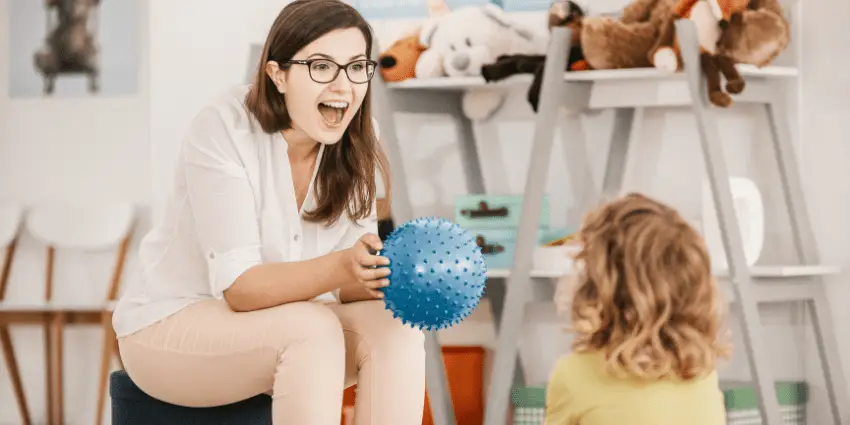 Speech Therapist engaging with a young child, holding a blue spiky ball during a therapy session in a room with shelves of toys.